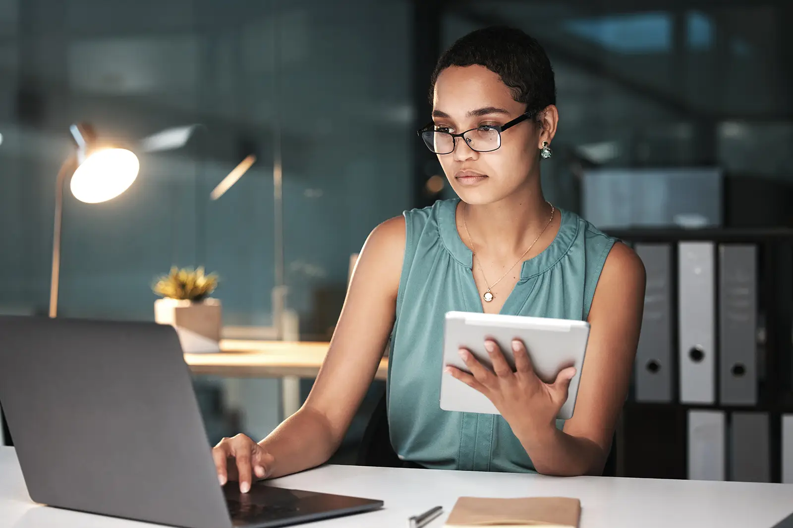 Eine Frau sitzt am Computer und schaut in den Bildschirm, dahinter steht eine Lampe